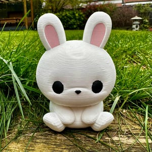 Puede incluir: Una figura de conejo blanco con grandes ojos negros y orejas internas rosas. El conejo est&aacute; sentado sobre una superficie de madera, rodeado de hierba verde. La figura tiene un dise&ntilde;o lindo y de dibujos animados.