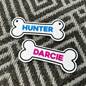 Puede incluir: Dos etiquetas blancas en forma de hueso para mascotas con borde negro. Una etiqueta tiene el nombre "HUNTER" en azul, y la otra "DARCIE" en rosa. Ambas etiquetas tienen un peque&ntilde;o agujero para sujetar.