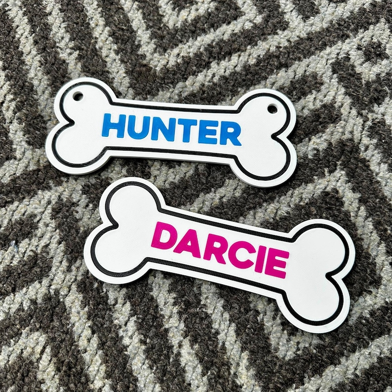 Puede incluir: Dos etiquetas blancas en forma de hueso para mascotas con borde negro. Una etiqueta tiene el nombre "HUNTER" en azul, y la otra "DARCIE" en rosa. Ambas etiquetas tienen un peque&ntilde;o agujero para sujetar.