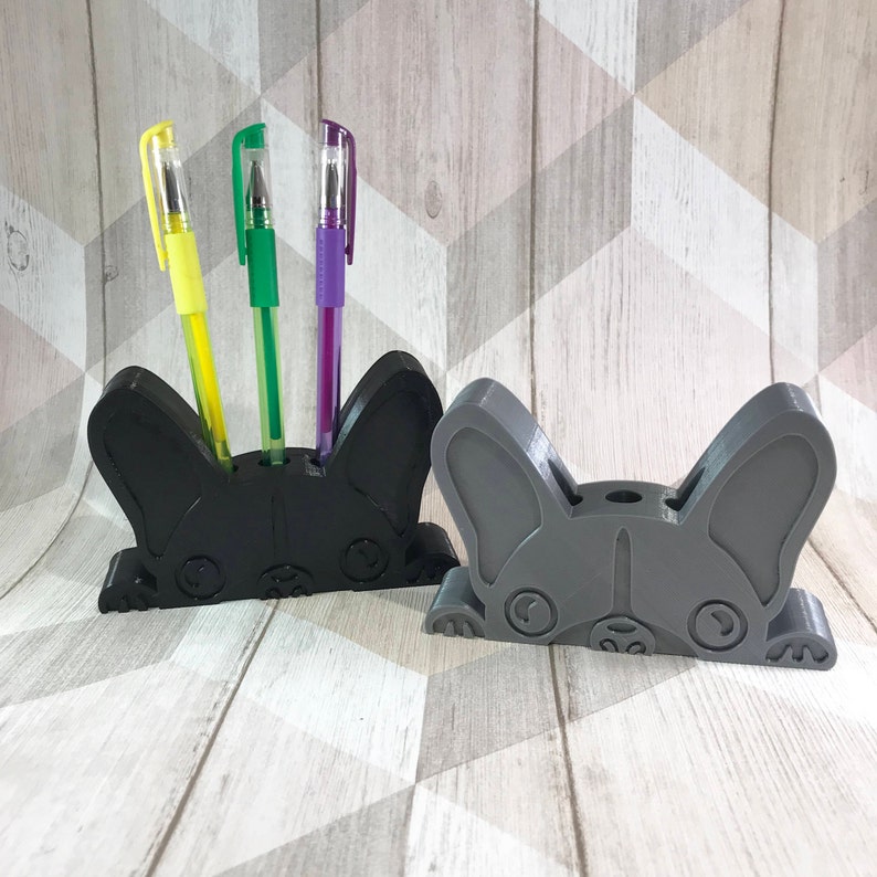 May include: Two 3D printed French Bulldog pencil holders. One is black and the other is gray. Both have a cute cartoon face and three holes for pencils. The black holder has three pens in it: one yellow, one green, and one purple.