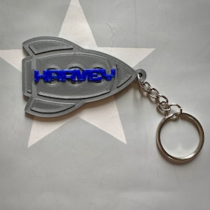 Peut inclure: Un porte-clés en forme de fusée imprimé en 3D de couleur grise avec le nom "HARVEY" imprimé en bleu. Le porte-clés est doté d'une chaîne en métal argenté et d'un anneau en métal argenté.
