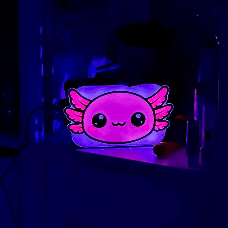 K&ouml;nnte beinhalten: Ein rosa Axolotl-Nachtlicht im Cartoon-Design mit gro&szlig;en Augen und federartigen Kiemen. Das rechteckige Licht strahlt ein sanftes violettes Leuchten aus, geeignet f&uuml;r ein Kinderzimmer. Ma&szlig;e nicht verf&uuml;gbar.