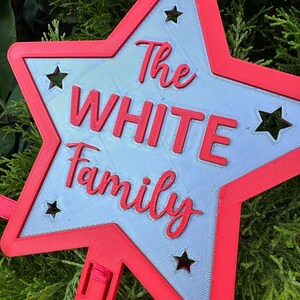 Puede incluir: Una decoraci&oacute;n en forma de estrella roja y gris con las palabras "The WHITE Family" en escritura roja. La estrella tiene formas de estrella recortadas y un borde rojo. La decoraci&oacute;n es probablemente para una familia.