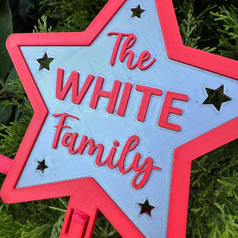 Puede incluir: Una decoraci&oacute;n en forma de estrella roja y gris con las palabras "The WHITE Family" en escritura roja. La estrella tiene formas de estrella recortadas y un borde rojo. La decoraci&oacute;n es probablemente para una familia.