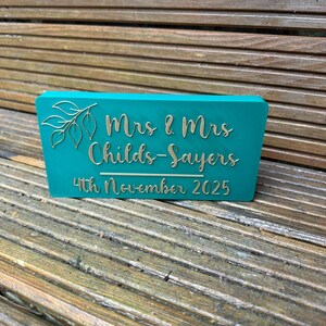 Op de afbeelding: Een turquoise rechthoekig bord met gouden letters en een bladontwerp. Op het bord staat "Mrs & Mrs Childs-Sayers 4th November 2025". Het bord staat op een houten oppervlak.