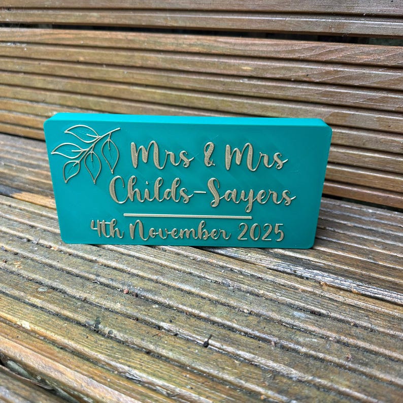 Op de afbeelding: Een turquoise rechthoekig bord met gouden letters en een bladontwerp. Op het bord staat "Mrs & Mrs Childs-Sayers 4th November 2025". Het bord staat op een houten oppervlak.