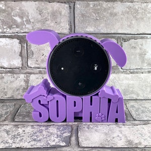 Puede incluir: Soporte de perro de color morado impreso en 3D con el nombre "Sophia" impreso en la base. El soporte contiene un Amazon Echo Dot negro.