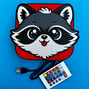 May include: A raccoon-shaped LED light with a red background and a white remote control. The raccoon has black and white fur, large eyes, and a pink tongue. The remote has multiple color options.