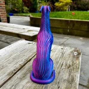 May include: A 3D-printed sculpture of a dog in a sitting position. The dog is primarily blue and purple, with vertical stripes of color. The sculpture has a long, slender body and a curved tail. The dog is sitting on a wooden surface.