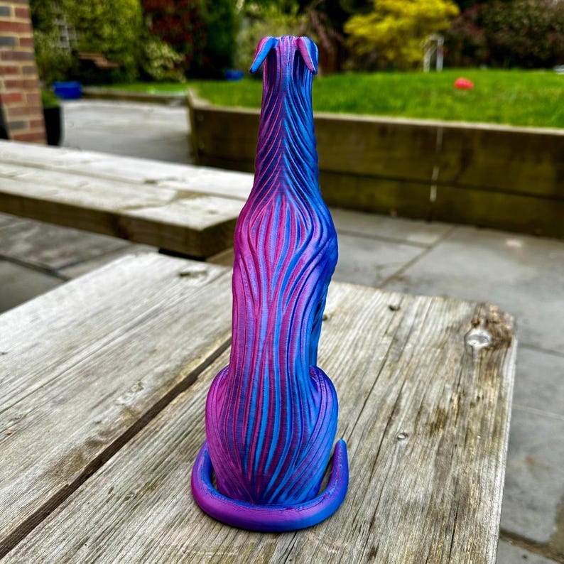 May include: A 3D-printed sculpture of a dog in a sitting position. The dog is primarily blue and purple, with vertical stripes of color. The sculpture has a long, slender body and a curved tail. The dog is sitting on a wooden surface.
