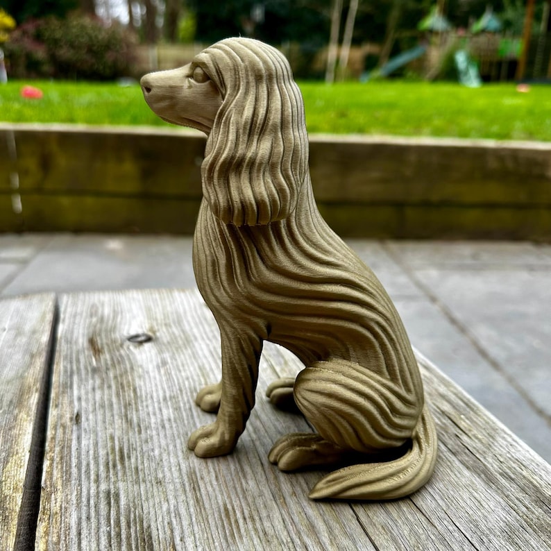 May include: A detailed, seated dog sculpture with a textured, wavy design. The dog is a light brown color and is sitting on a wooden surface. The sculpture has long, flowing hair and is facing left.