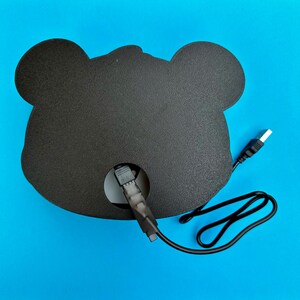 May include: A black, bear-shaped LED light with a USB cable for power. The light has a circular opening in the center and is set against a bright blue background. The cable is black with a white USB connector.