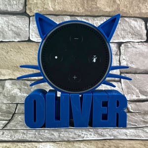 Puede incluir: Un soporte para altavoz inteligente Amazon Echo Dot con forma de gato impreso en 3D de color azul. El soporte tiene el nombre "OLIVER" impreso en la parte delantera.
