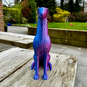 May include: A decorative dog figurine in shades of blue and purple, sitting upright. The dog has a long neck and detailed fur-like texture. The figurine is placed on a weathered wooden surface, with a blurred outdoor background.