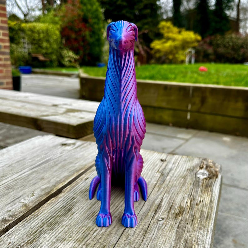 May include: A decorative dog figurine in shades of blue and purple, sitting upright. The dog has a long neck and detailed fur-like texture. The figurine is placed on a weathered wooden surface, with a blurred outdoor background.