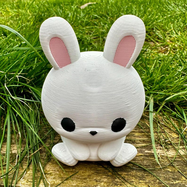 Puede incluir: Una figura de conejo blanco con grandes ojos negros y el interior de las orejas rosa. El conejo est&aacute; sentado con las patas hacia adelante y tiene orejas largas y erguidas. La figura es de material texturizado, sobre hierba verde.