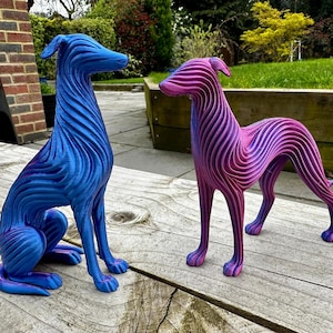 May include: Two decorative dog sculptures. One is blue with purple accents, the other is pink and purple. Both have a striped texture and are posed on a wooden surface. The sculptures are in a garden setting.