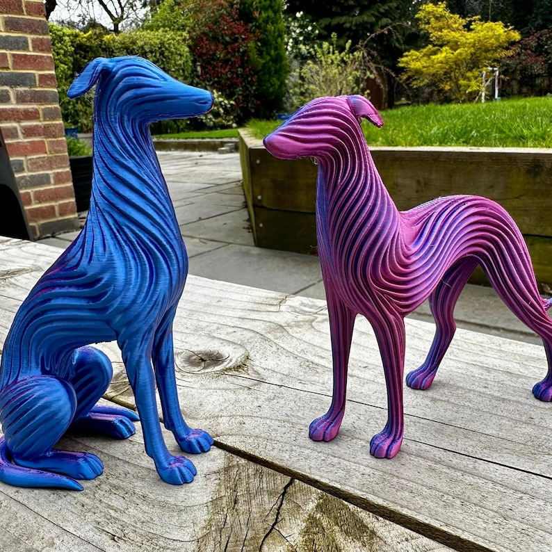 May include: Two decorative dog sculptures. One is blue with purple accents, the other is pink and purple. Both have a striped texture and are posed on a wooden surface. The sculptures are in a garden setting.