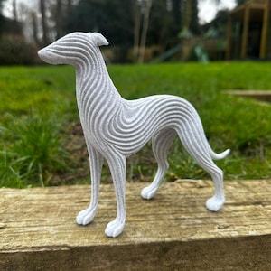 May include: A gray, stylized sculpture of a dog, possibly a greyhound, with a textured, layered design. The dog is standing on a wooden surface, with a green lawn and trees in the background. The sculpture is a decorative item.
