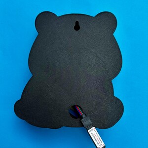 May include: Black bear-shaped wall decor with a textured surface. The back features a keyhole for hanging and a multi-colored LED controller cable. The background is a solid blue.