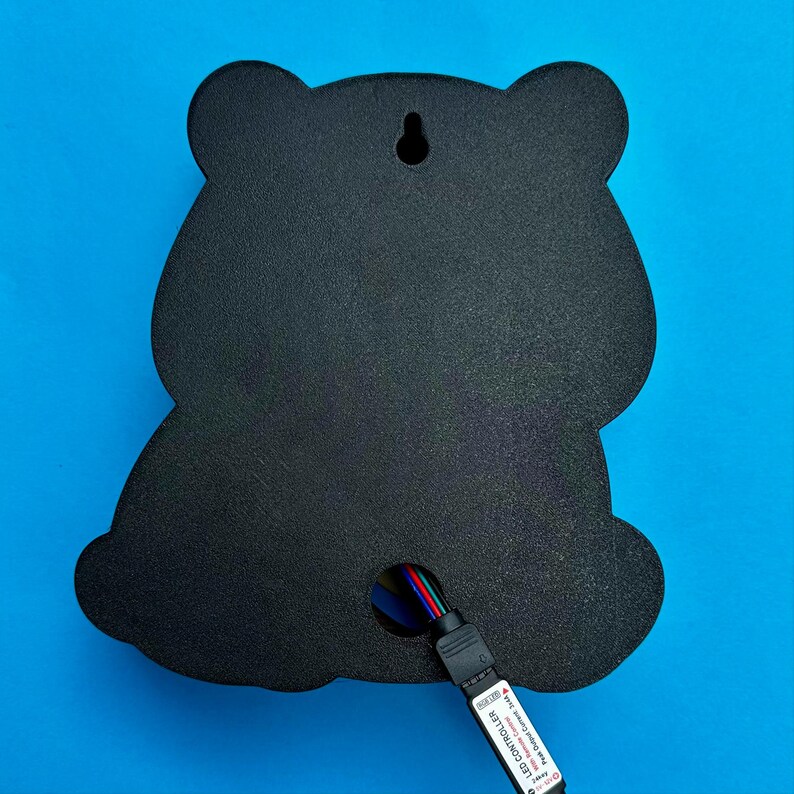 May include: Black bear-shaped wall decor with a textured surface. The back features a keyhole for hanging and a multi-colored LED controller cable. The background is a solid blue.