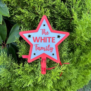 Puede incluir: Un letrero en forma de estrella roja y blanca con el texto "The WHITE Family". El letrero tiene un borde rojo, peque&ntilde;as estrellas negras y un clip rojo en la parte inferior. Decoraci&oacute;n de jard&iacute;n.