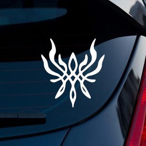 May include: A white vinyl decal featuring a symmetrical, abstract, flame-like or wing-like design with intricate, flowing lines that curve upward and outward from a central point, applied to the rear window of a dark-colored vehicle.