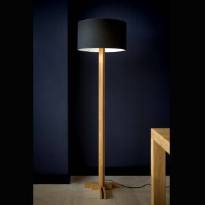 Op de afbeelding: Een houten vloerlamp met een zwarte stoffen kap. De lamp heeft een eenvoudig, modern ontwerp en is perfect om elke kamer een vleugje stijl te geven.