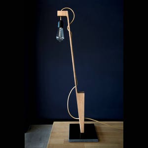 Op de afbeelding: Een houten vloerlamp met een zwart snoer en een gloeilamp. De lamp heeft een uniek, hoekig ontwerp en is gemaakt van natuurlijk hout.