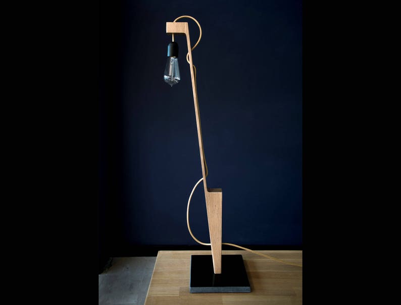 Op de afbeelding: Een houten vloerlamp met een zwart snoer en een gloeilamp. De lamp heeft een uniek, hoekig ontwerp en is gemaakt van natuurlijk hout.