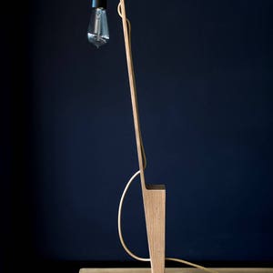 Op de afbeelding: Een houten vloerlamp met een zwart snoer en een gloeilamp. De lamp heeft een eenvoudig, modern ontwerp en is gemaakt van natuurlijk hout.