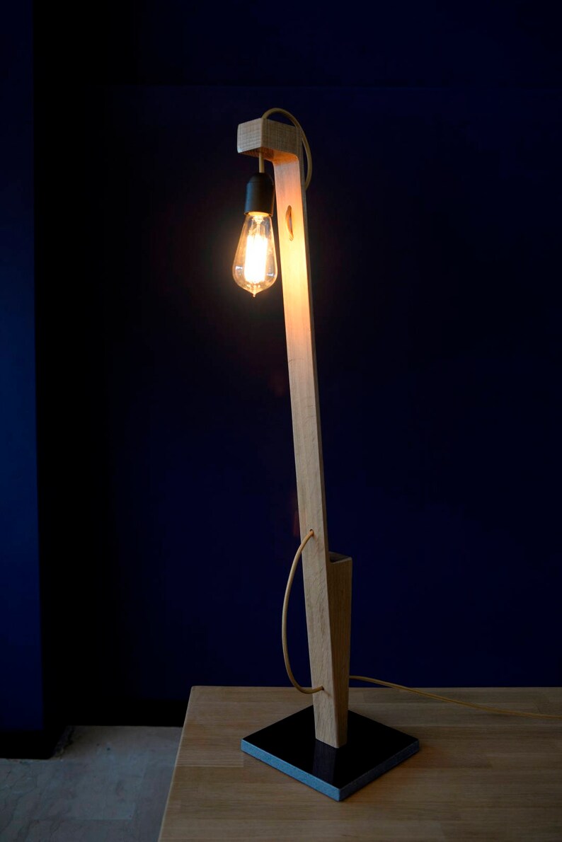 Op de afbeelding: Een houten vloerlamp met een zwarte voet en een gloeilamp. De lamp staat op een houten tafel.