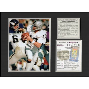 May include: A black and white photo of a football player in a white jersey with the number 12 on it. He is running with the ball and being tackled by a player in a purple jersey with the number 6 on it. The photo is from Super Bowl XI, where the Oakland Raiders defeated the Minnesota Vikings 32-14. The text "RAIDERS 32 VIKINGS 14" is visible in the image.