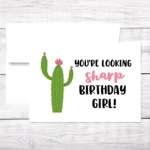 May include: A white greeting card with a green cactus illustration and the text "YOU'RE LOOKING sharp BIRTHDAY GIRL!" in black and pink. The card is paired with a white envelope.