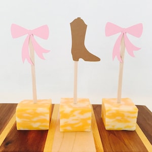 May include: Three cheese cubes on wooden skewers with pink bows and a brown boot cutout on top. The skewers are on a wooden cutting board with alternating brown and light wood stripes.
