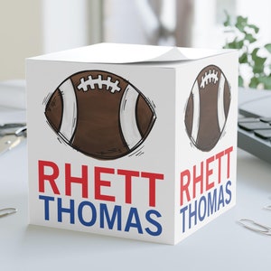 May include: White cube-shaped desk organizer with a football graphic and the name "Rhett Thomas" printed on two sides in red and blue lettering.