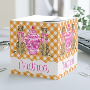 May include: A cube-shaped notepad with an orange and white gingham pattern. The design features cheetahs wearing pink bows and pink decorative vases. The name "Andrea" is written in pink script across the front.