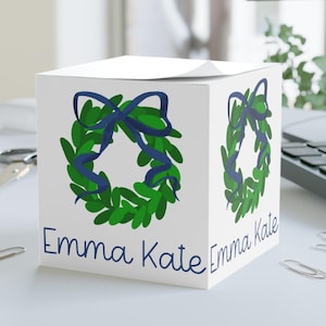 May include: White cube-shaped box with a green and blue wreath design and the text "Emma Kate" on the front.
