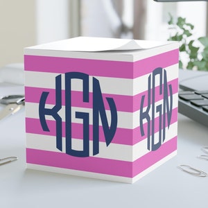 May include: A pink and white striped notepad with a navy blue monogram "KGN" on the front. The notepad is sitting on a white surface.