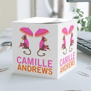 May include: White cube-shaped notepad with a repeating pattern of cartoon monkeys holding pink umbrellas. The text "CAMILLE ANDREWS" is printed in pink and orange. The notepad is on a white desk with paper clips.