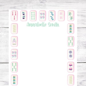 May include: A white notepad with a pink border features a design of mahjong tiles with various symbols and images. The name "Annabelle Smith" is printed at the top of the notepad.