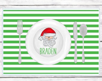 May include: Green and white striped placemat with a white plate featuring a cartoon Santa Claus with the name "Braden" printed below. A fork, knife, and spoon are placed on the placemat.
