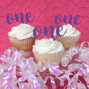 May include: Three cupcakes with purple 'one' toppers on a pink background with white iridescent ribbon.