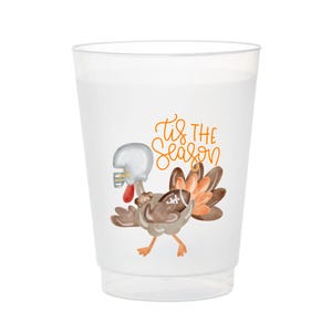 May include: A translucent plastic cup featuring a graphic of a turkey wearing a silver football helmet and holding a football. Above the turkey, the orange text reads 'tis THE season'. The cup has a tapered shape with a smooth, clear finish.