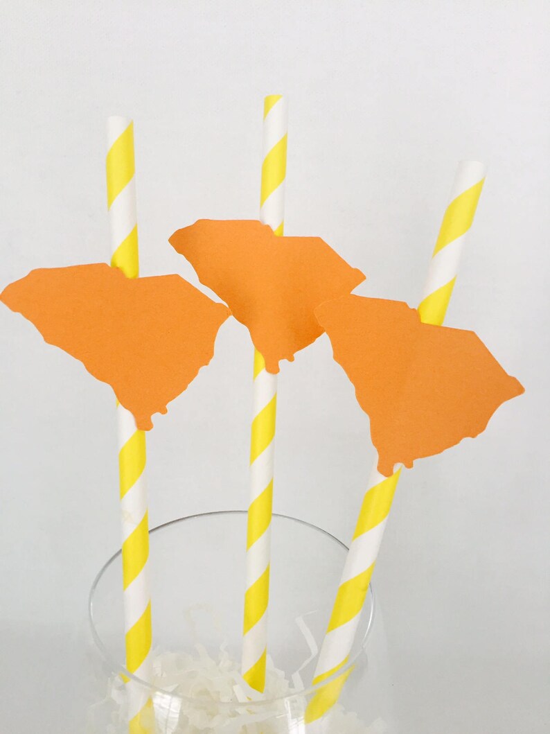 Pode incluir: Um conjunto de palhinhas de papel &agrave;s riscas amarelas e brancas, cada uma encimada por um recorte laranja do estado da Carolina do Sul. As palhinhas est&atilde;o dispostas num copo transparente, sugerindo um tema festivo.