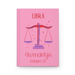 Pode incluir: Caderno rosa com o signo do zodíaco Libra, uma balança com dois pratos, em vermelho e roxo. O texto "LIBRA" está no topo do caderno. O nome "Gwendolyn" e a data "October 20" estão escritos abaixo da balança.
