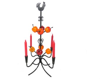 Puede incluir: Un candelabro de metal negro con un remate de gallo y velas rojas. El candelabro está decorado con fruta artificial en tonos naranja y rojo.