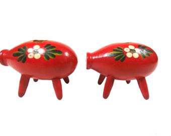 Puede incluir: Dos objetos decorativos rojos de madera, cada uno con tres patas y un cuerpo redondeado. Los objetos están pintados con flores blancas y hojas verdes. Los objetos tienen un agujero en un extremo.