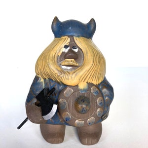 May include: A ceramic figurine of a Viking warrior with a blue hat, yellow hair, and a brown body. The warrior is holding an axe and a shield.