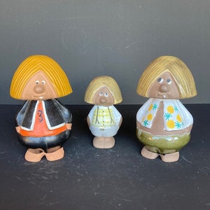 May include: Three ceramic figurines of people with blonde hair and painted faces. The figurine on the left is wearing a black jacket with an orange collar and the number "3" on the front. The middle figurine is wearing a white shirt with a yellow and white checked pattern. The figurine on the right is wearing a brown shirt with a yellow and green floral pattern.